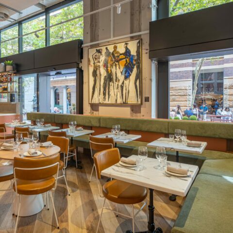 Margot Osteria's dining area features a soft palette, fresh finishes, and refined textures. Olive green banquette seating wraps the interior with leather-upholstered chairs and sleek stone tabletops, offering comfort and contrast against the building’s raw structural shell.